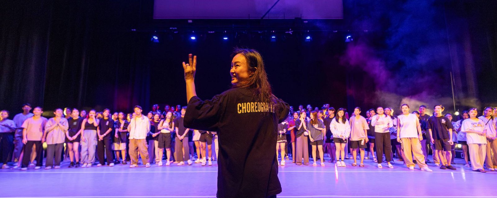 Stephanie Loh in a choreographer shirt waving to a large group of dancers on a purple-lit stage